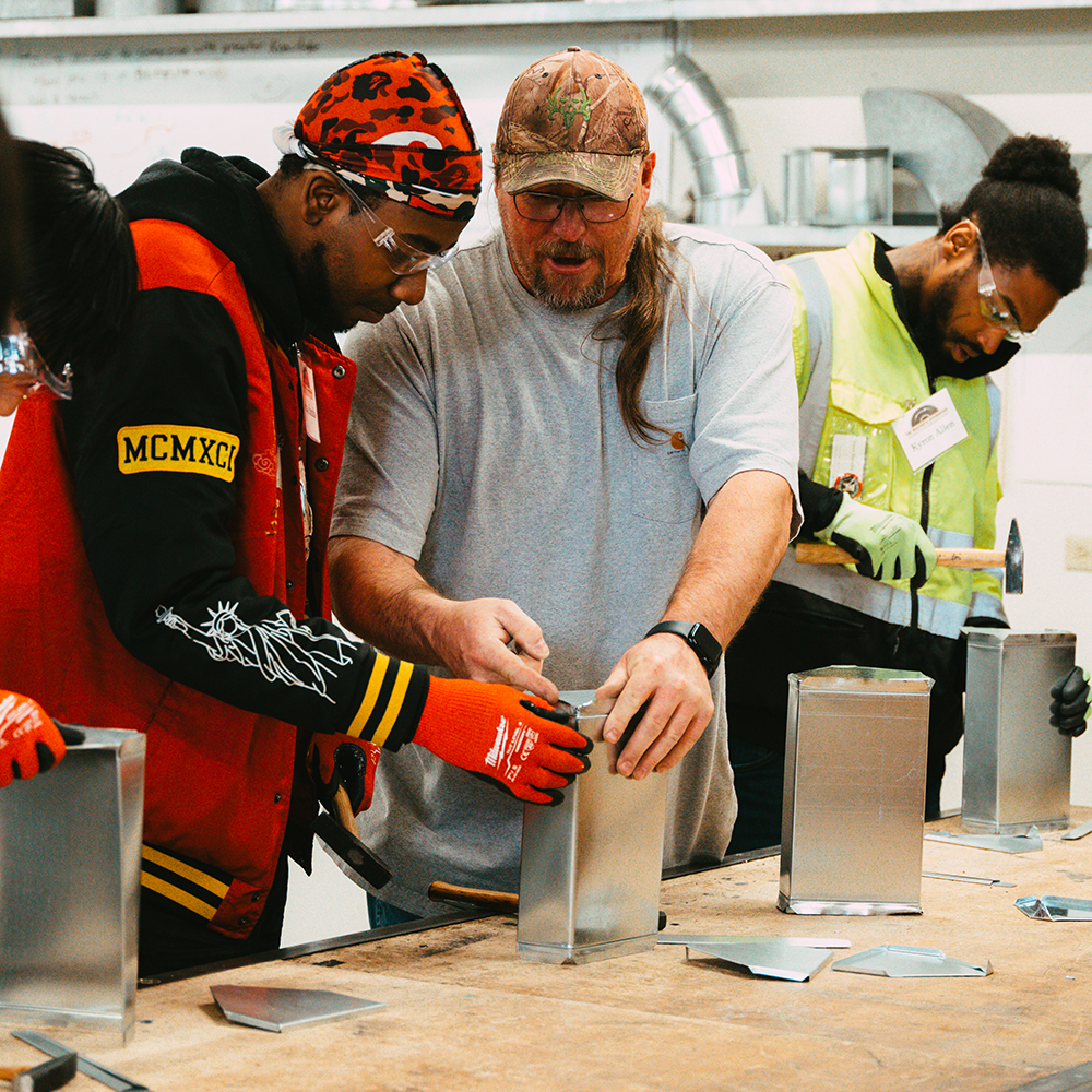 Instructor teaches two students how to work with metal sheets on a table in a workshop.